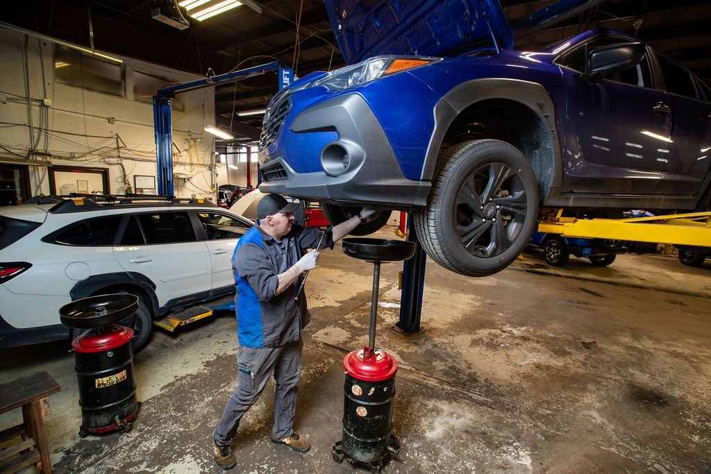 Technician inspecting a blue Subaru on a lift in the Juneau Auto Mall service shop