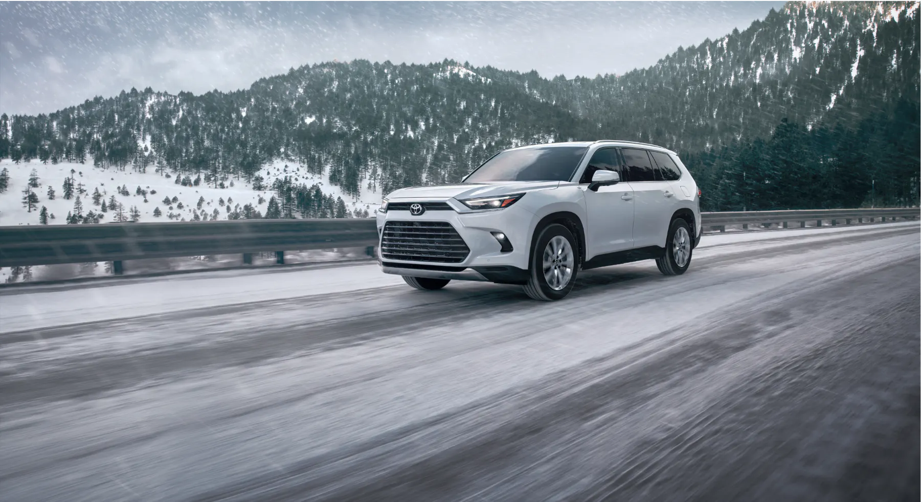 White Toyota Grand Highlander family SUV driving on a snowy road with snow-covered mountains in the background