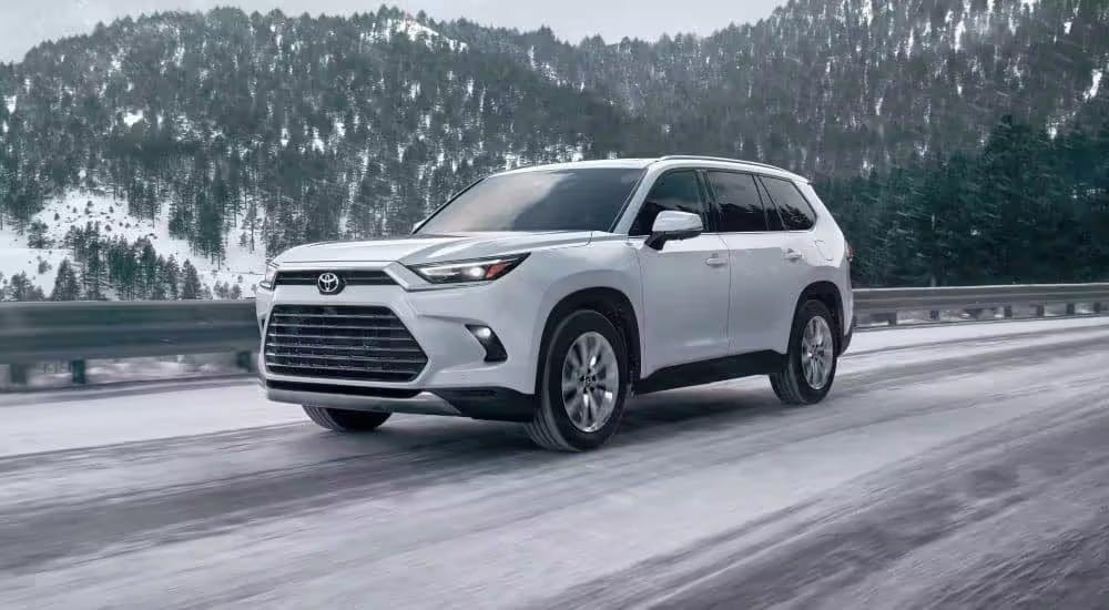 A white 2024 Toyota Grand Highlander is shown driving down a snowy road.