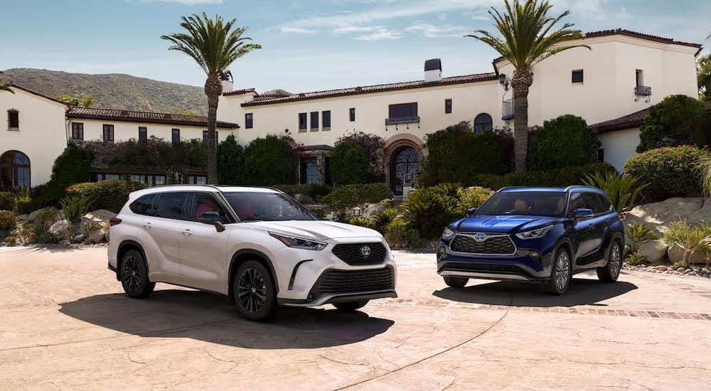 A white 2024 Toyota Highlander XSE and a blue 2024 Highlander Hybrid Platinum parked outside a villa.