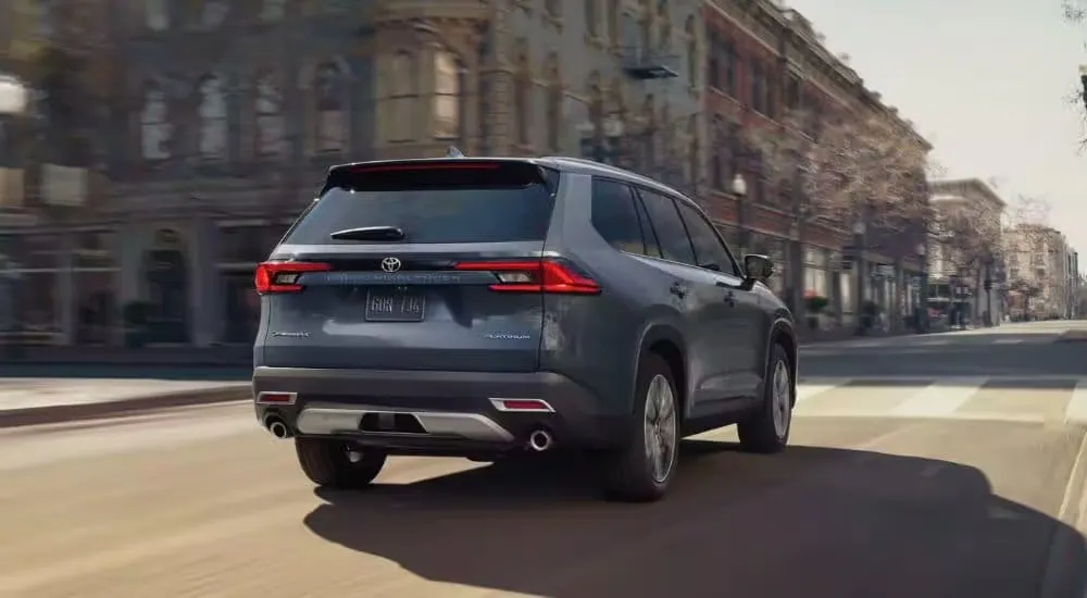 A grey 2024 Toyota Grand Highlander Platinum is shown from the rear driving on a city street.