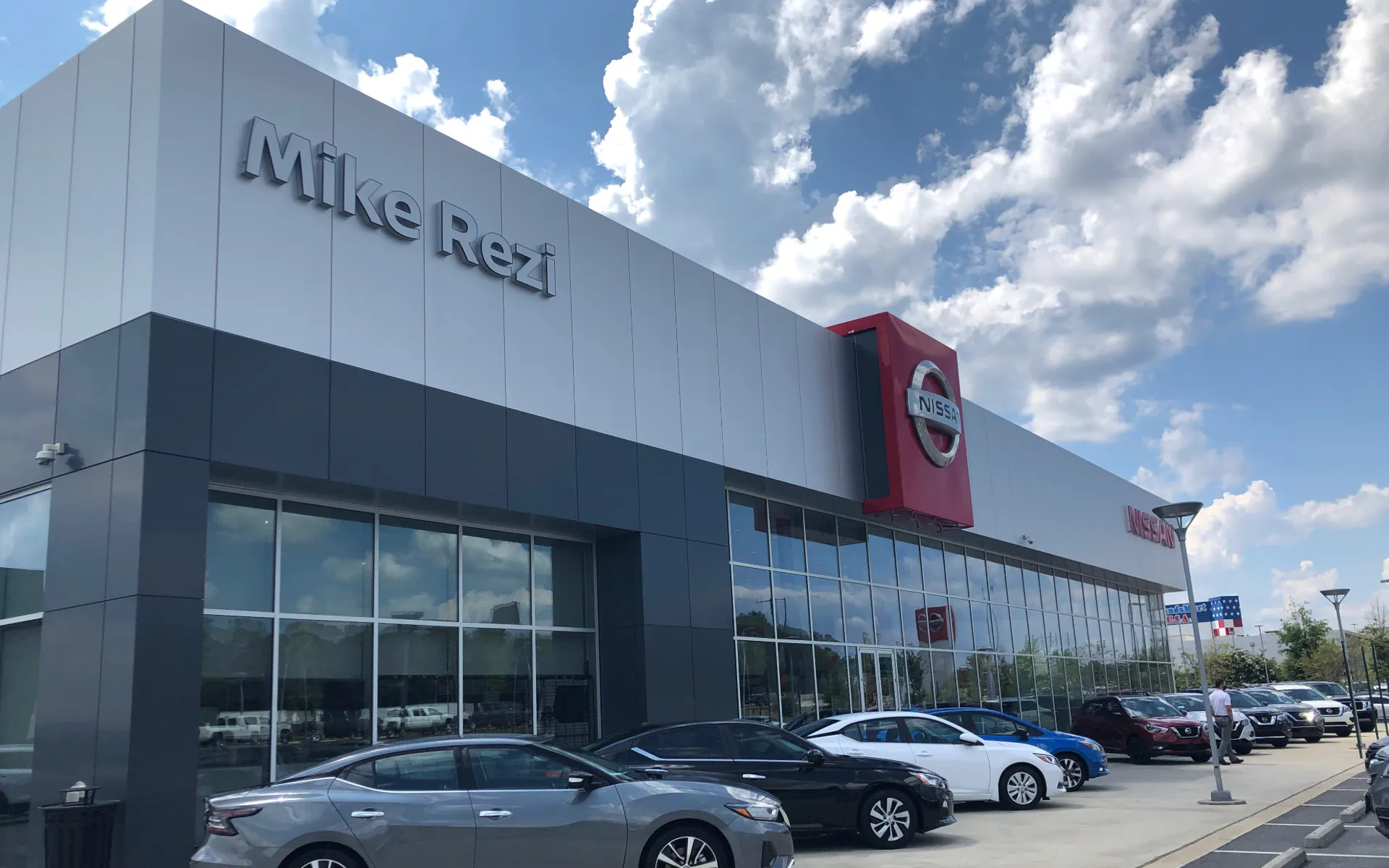 Exterior view of Mike Rezi Nissan dealership showroom with parked vehicles under a bright sky with scattered clouds.