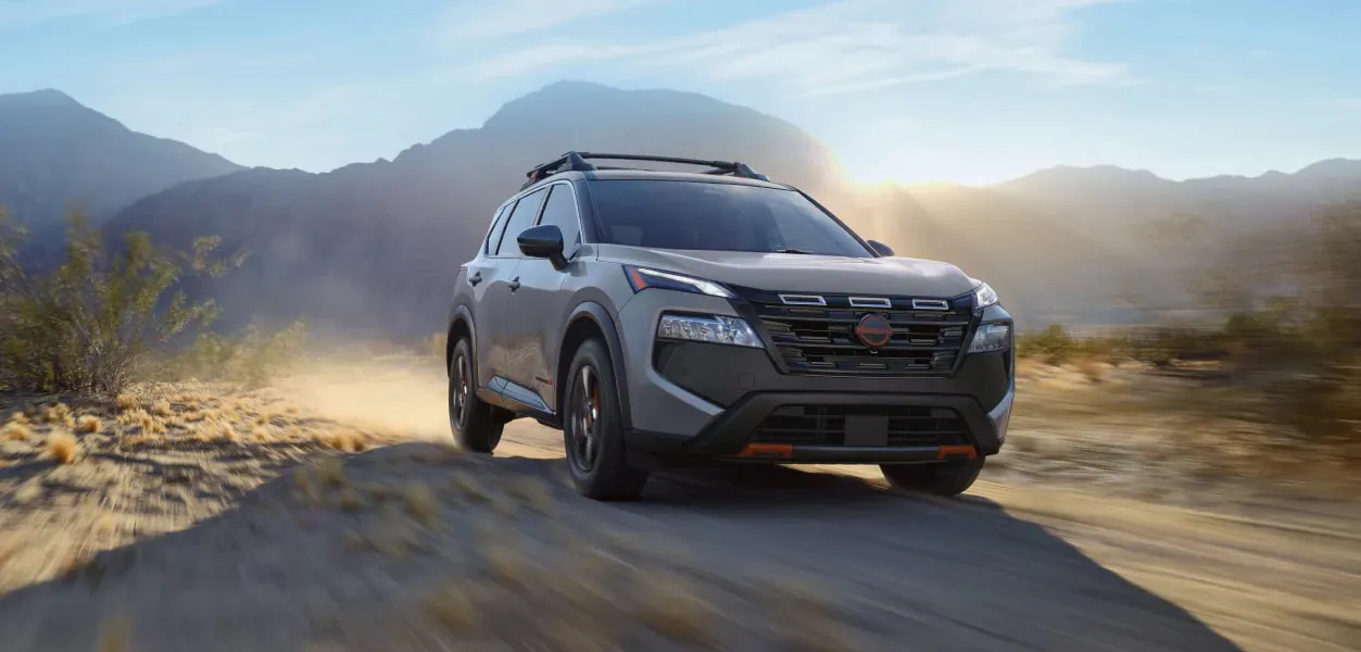 Gray Nissan SUV driving on a dusty desert trail with mountains in the background at sunset.