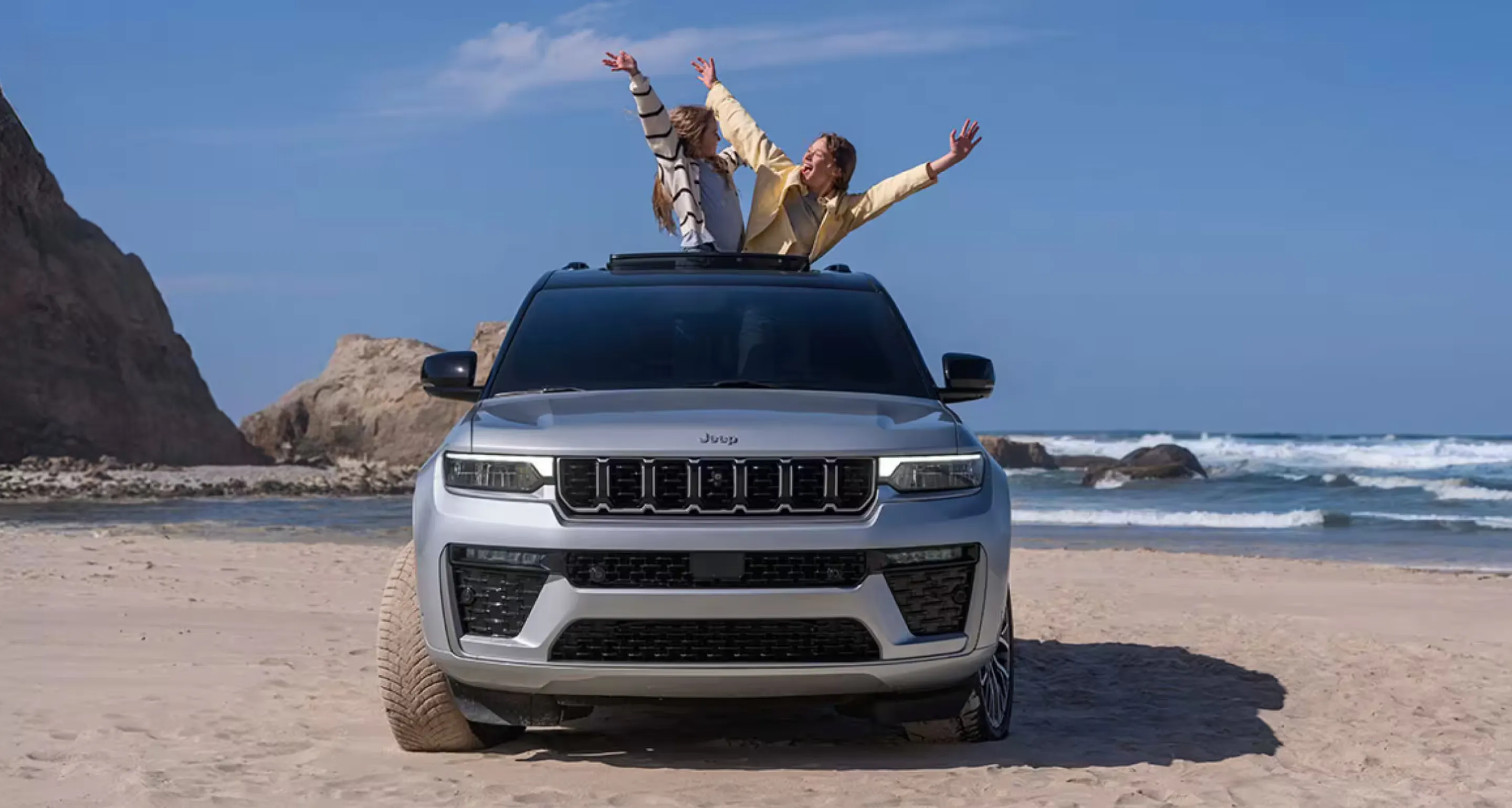 Silver Jeep SUV parked on a beach with two people standing through the open sunroof and raising their arms.