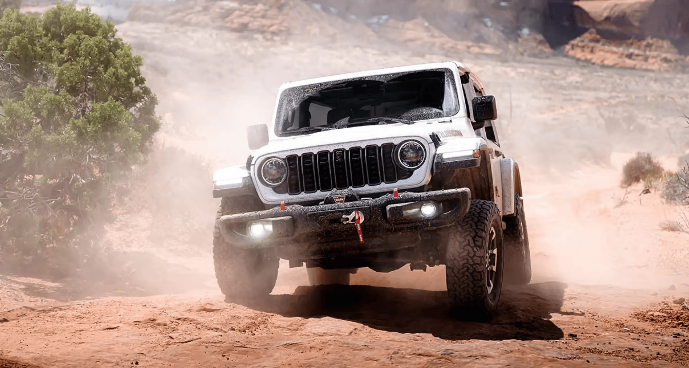 White Jeep off-road vehicle driving across a dusty rocky trail with its headlights on.