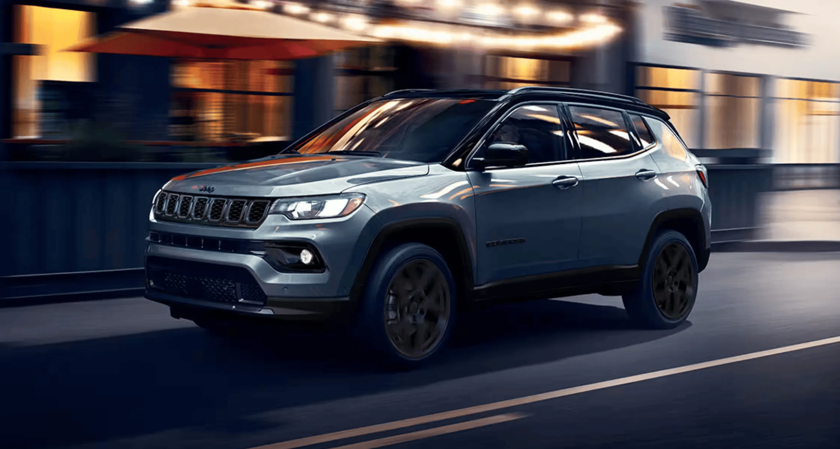 Gray Jeep SUV driving along a city street at night with motion-blurred buildings in the background.