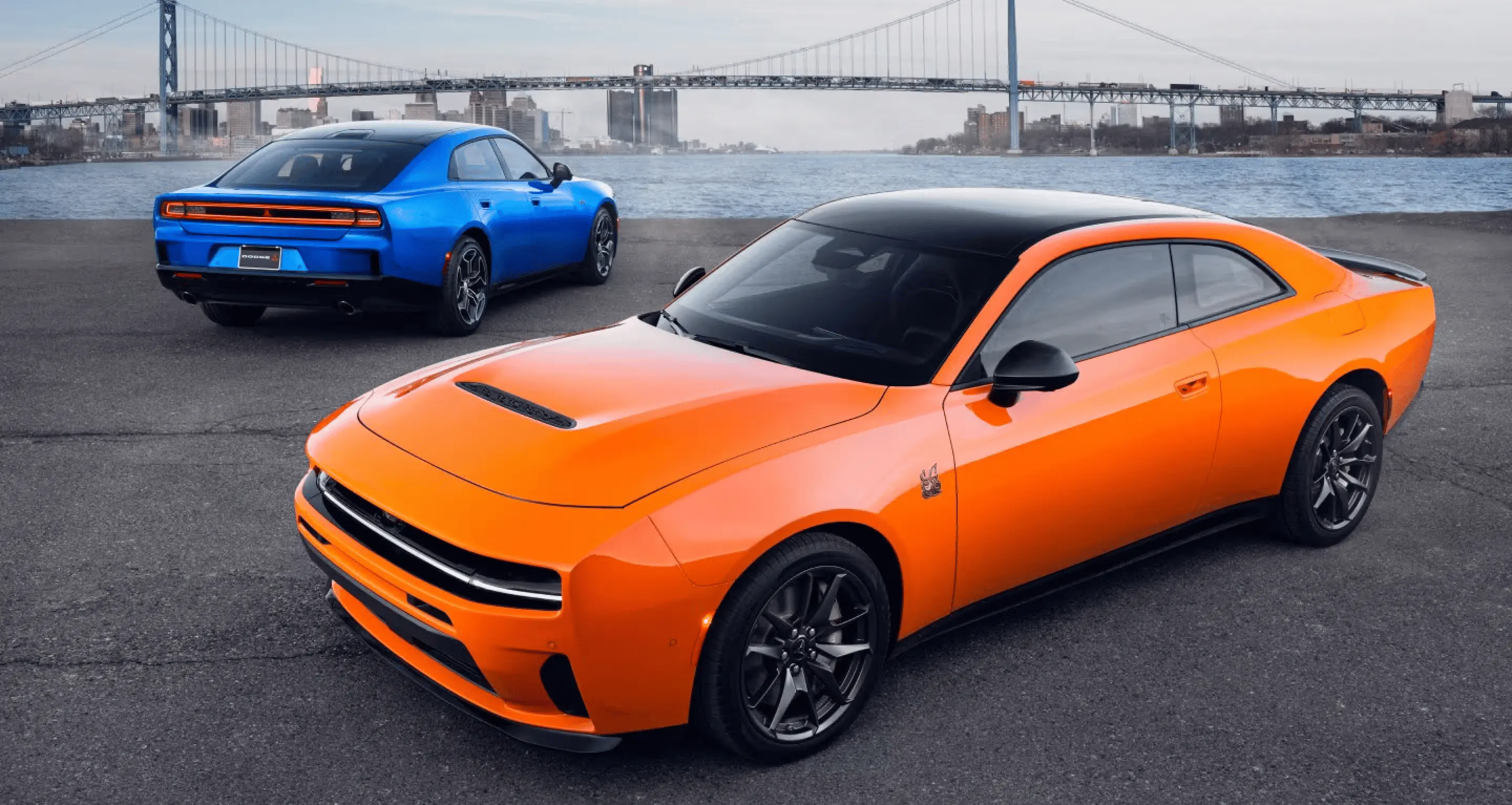 Orange sports coupe in the foreground and blue sedan behind it beside a river with a suspension bridge and city skyline.