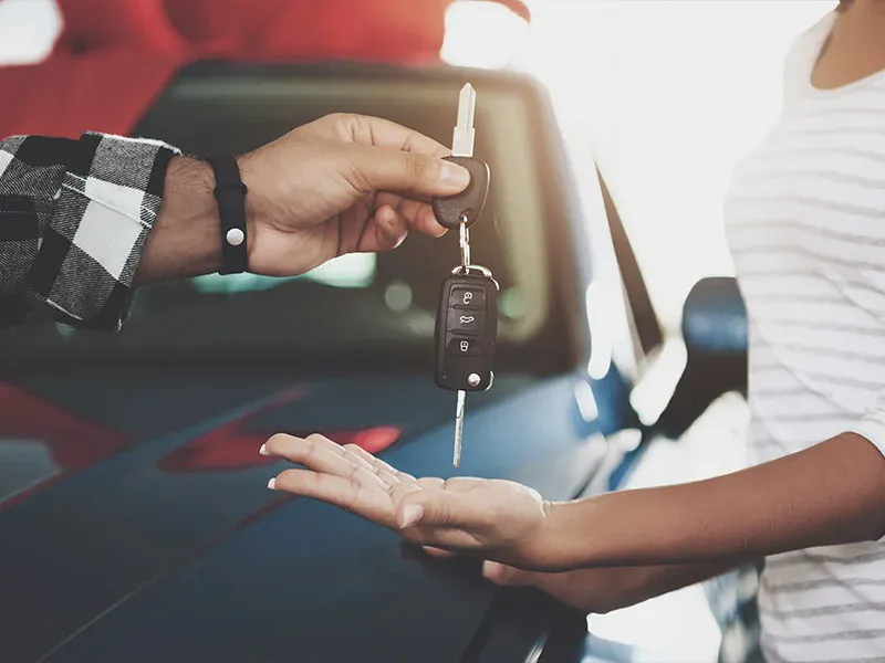 Person handing over car keys to a customer beside a vehicle, symbolizing vehicle purchase, trade-in, or rental delivery.