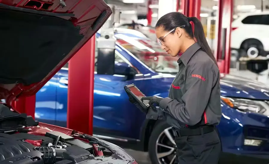 nissan-express-service-multi-point-1 Nissan service technician using a diagnostic tablet while inspecting a vehicle with the hood open inside an automotive service center.