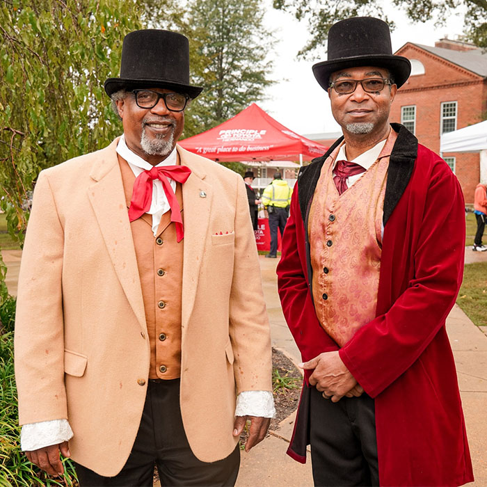Two distinguished gentlemen in top hats pose for a picture with event tent in background