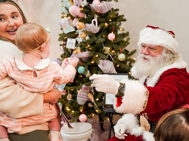 Santa seating in front of tree reaches out to a child being held by mother