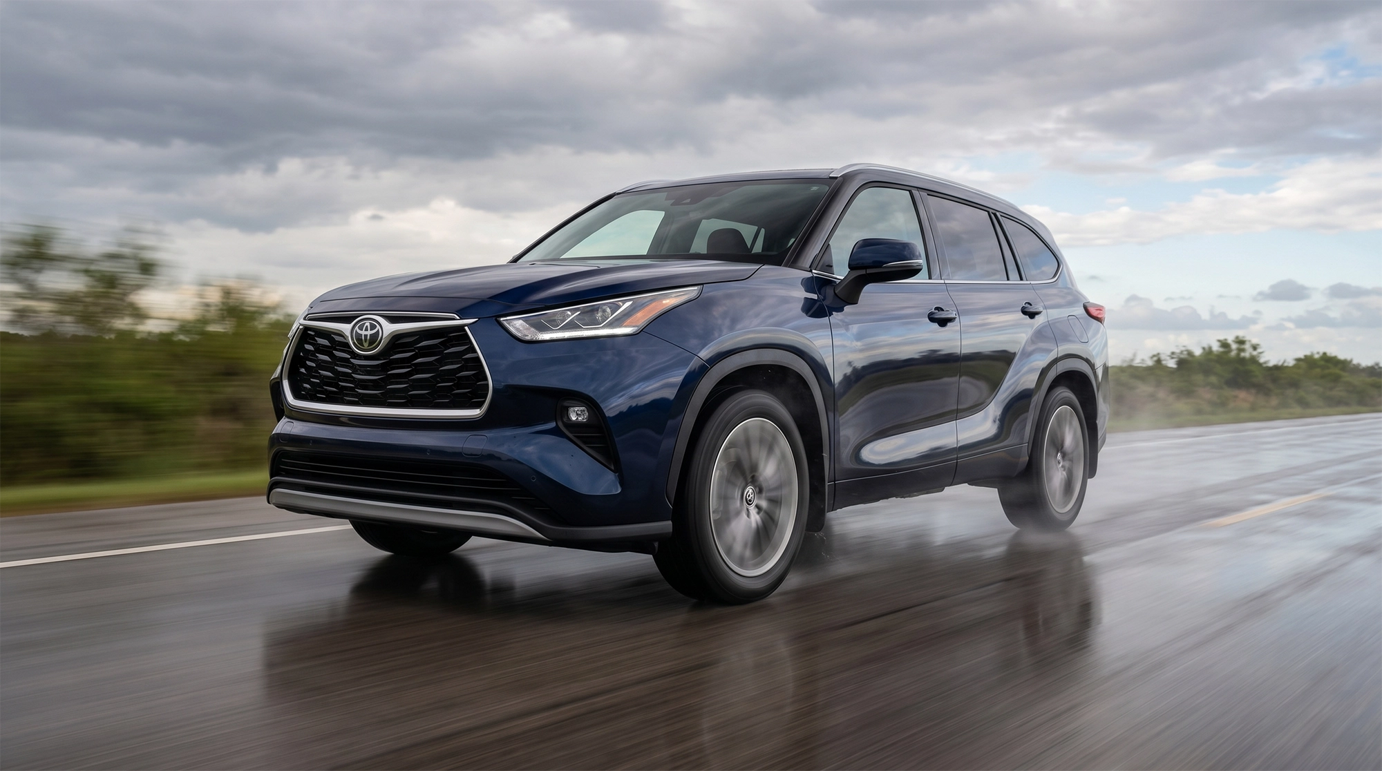 2026 Toyota Highlander in blue driving on a wet road in rainy conditions with water spray and cloudy sky