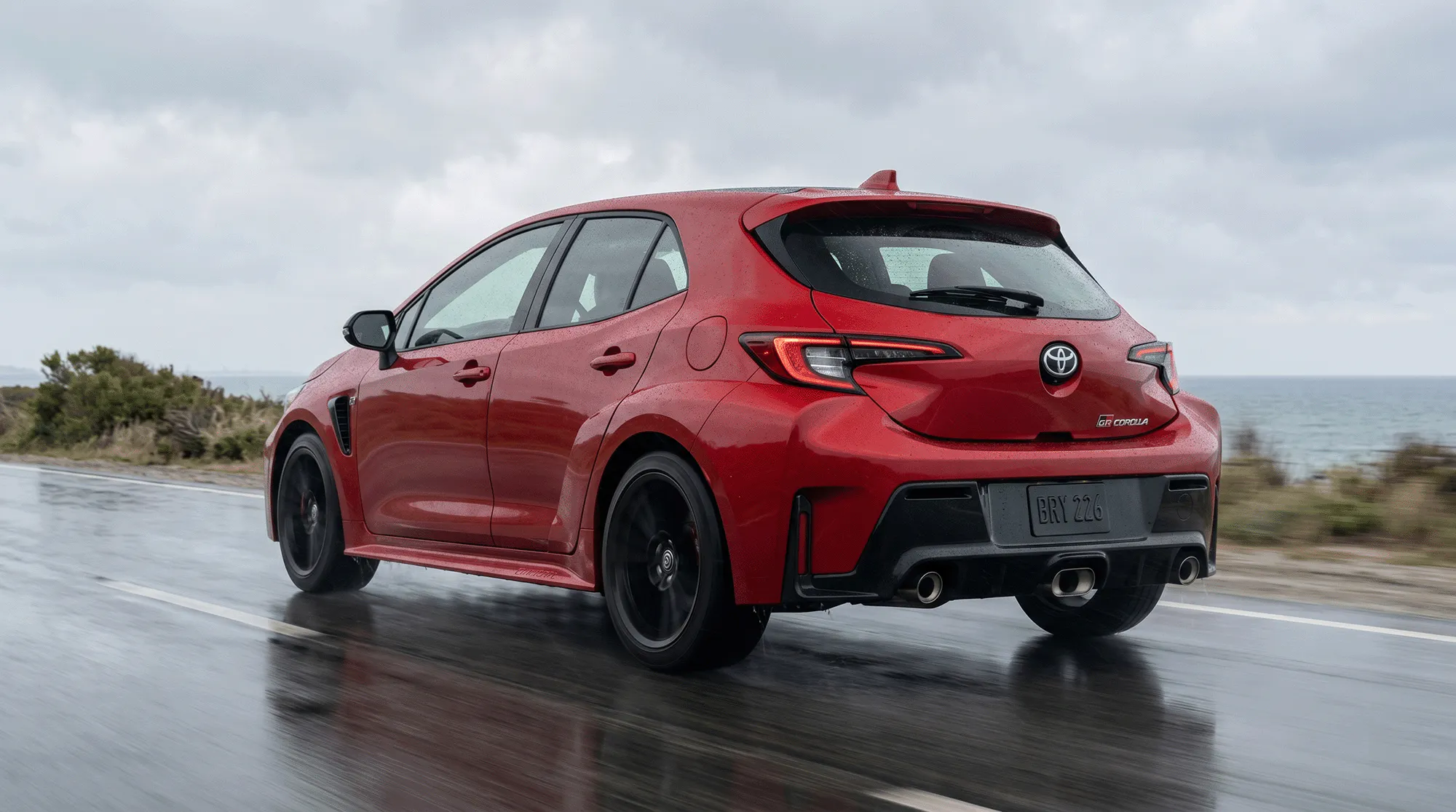 Red 2026 Toyota GR Corolla parked on a rain-soaked coastal road lined with palm trees, shown from the front three-quarter angle under dark storm clouds.