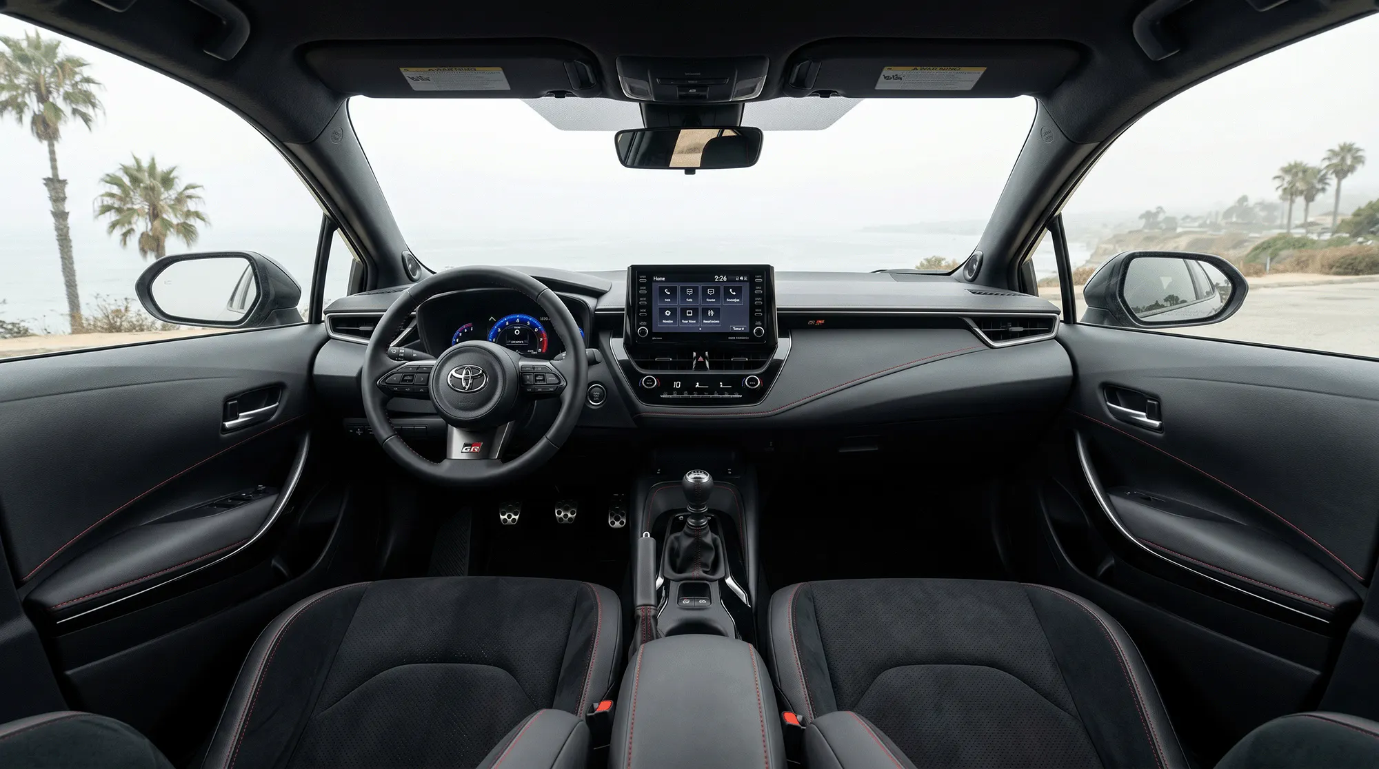 Interior view of the 2026 Toyota GR Corolla showing the dashboard, steering wheel, manual shifter, infotainment screen, and front seats with a coastal view through the windshield.