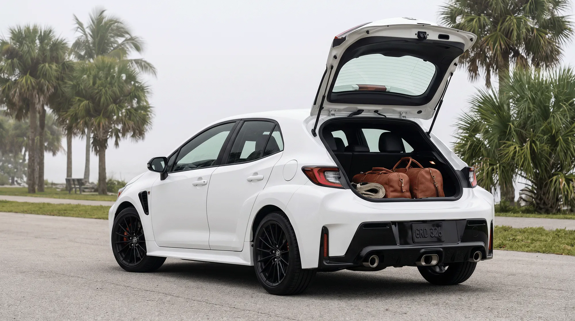 White 2026 Toyota GR Corolla with the rear hatch open, showing luggage in the cargo area beside palm trees in a coastal setting.