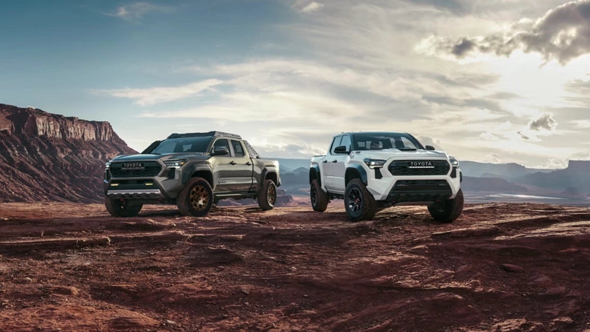 Two 2025 Toyota Tacoma i-Force Max trucks, one gray and one white, parked on rugged red rock terrain with desert cliffs and a cloudy sky in the background.