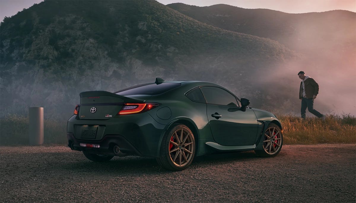 25-09-25_SPTOY_GR86 2025 Toyota GR86 sports car in dark green parked on a scenic mountain road at sunset, showcasing its sleek coupe design with bronze wheels and rear spoiler.
