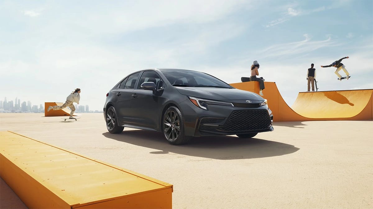 2025 Toyota Corolla in sleek dark gray parked near a skatepark with skaters performing tricks in the background on a sunny day.