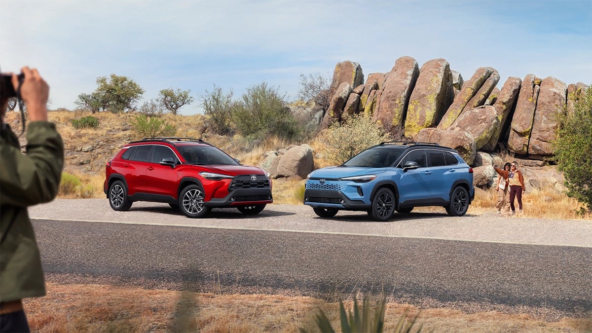 2025 Toyota Corolla Cross Hybrid in red and blue parked on a scenic desert road with rocky formations in the background, highlighting its modern SUV design.