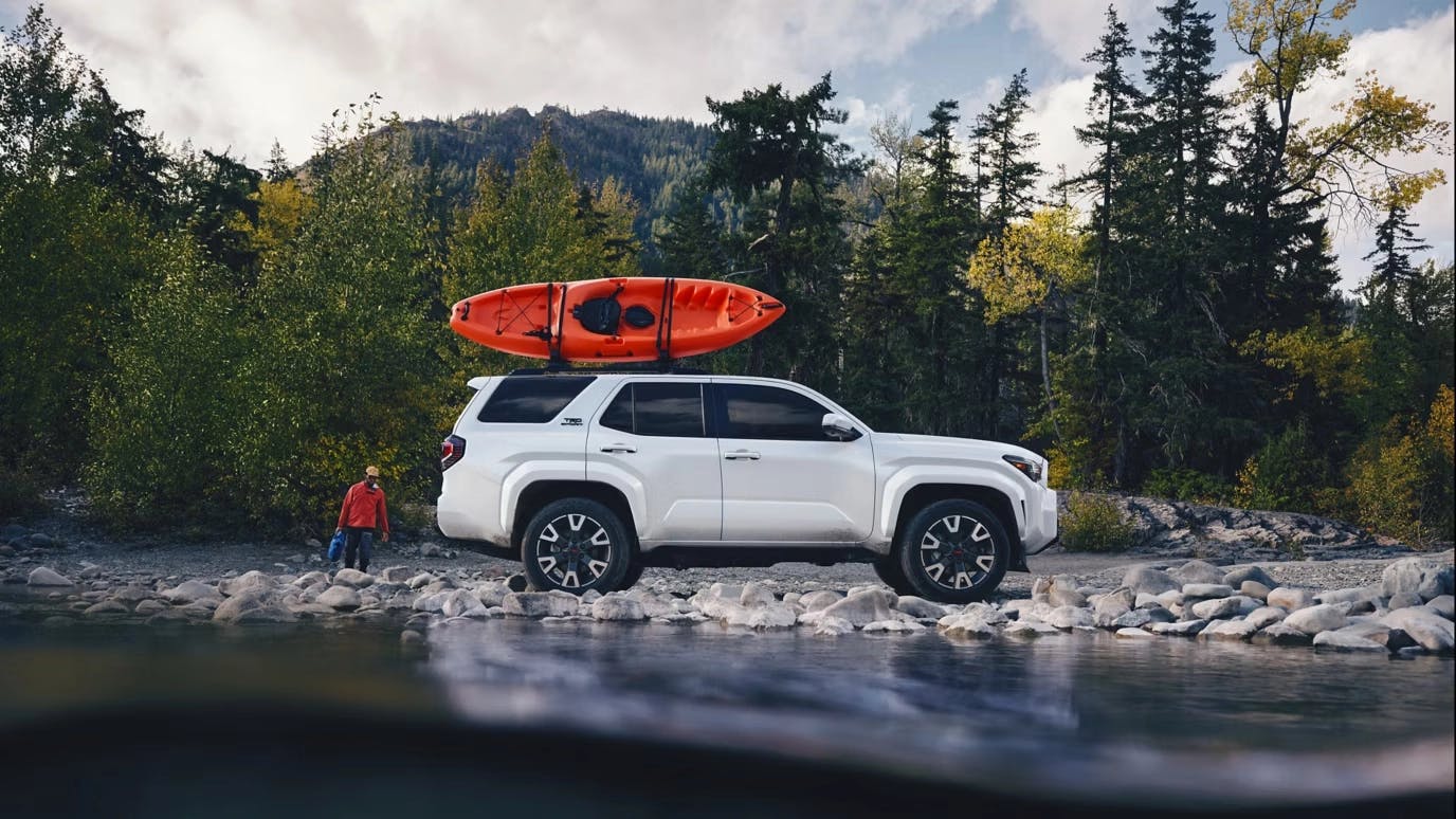 25-09-29_Toyota_SPTOY_4Runner i-Force MAX White Toyota 4Runner i-Force Max SUV parked by a river with an orange kayak mounted on the roof, surrounded by trees and mountains, showcasing its adventure-ready capability.