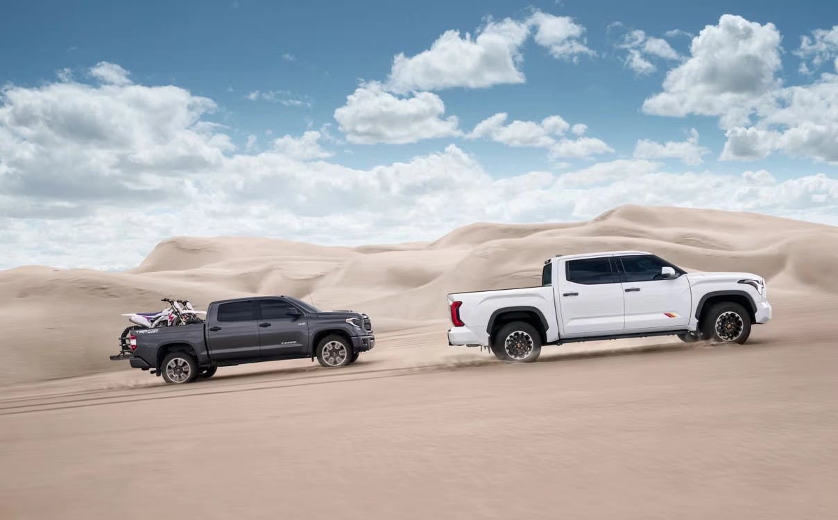 Two Toyota Tundra i-Force Max trucks driving across sandy dunes under a bright blue sky, showcasing off-road capability and towing power, with one truck carrying dirt bikes in the bed.