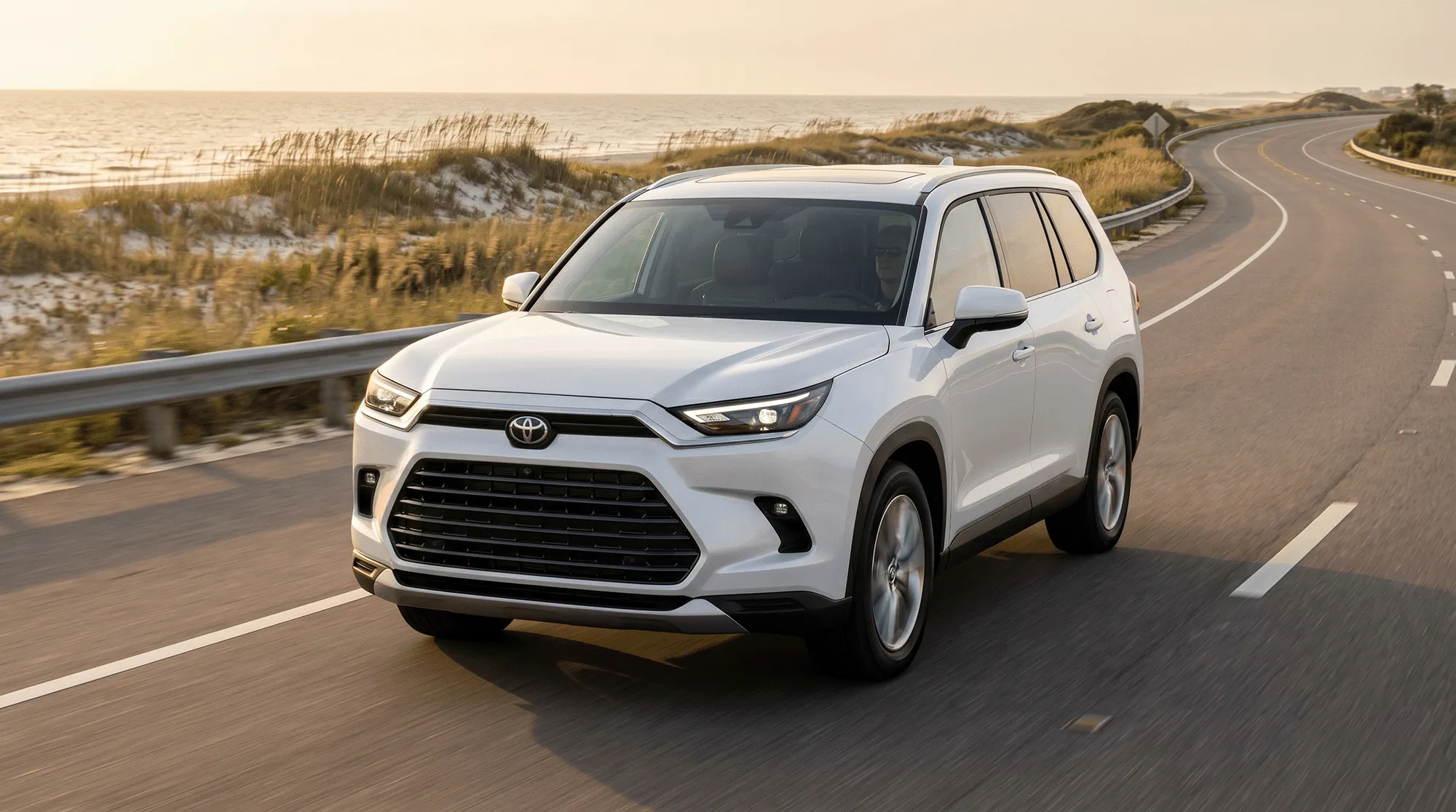 White 2026 Toyota Grand Highlander Hybrid driving along a scenic Gulf Coast road at golden hour